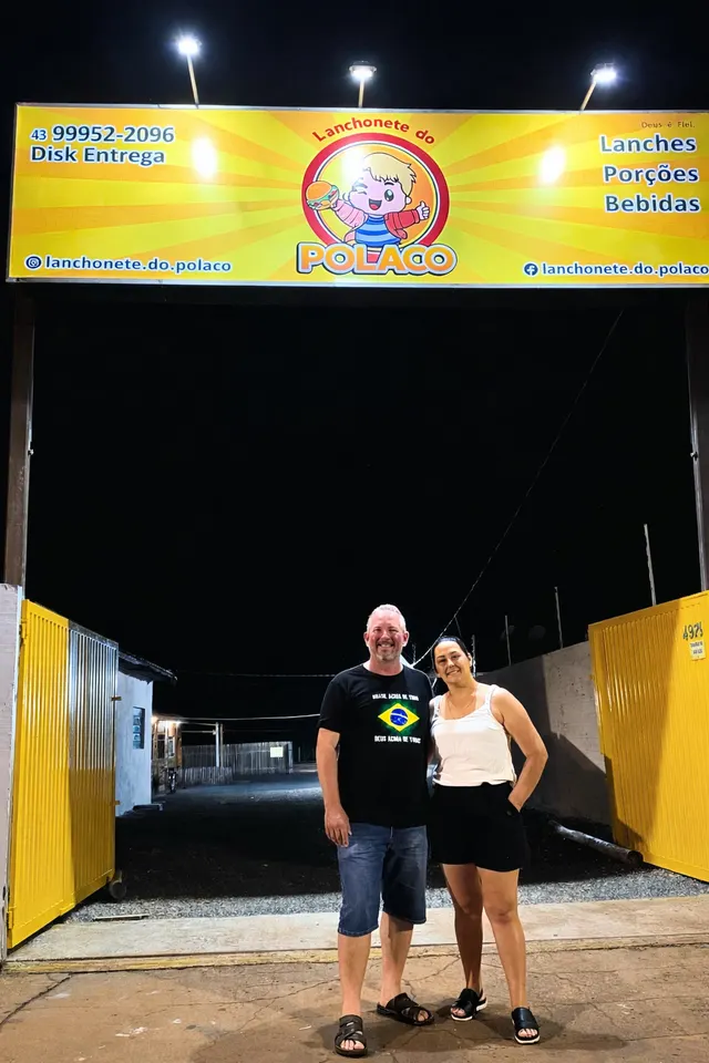 Michael, o Polaco, e Josiane no portão da Lanchonete do Polaco, à noite, sob a placa amarela